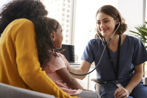 Female Registered Nurse RN Wearing Scrubs Listening To Girls Chest With Stethoscope In Hospital Office